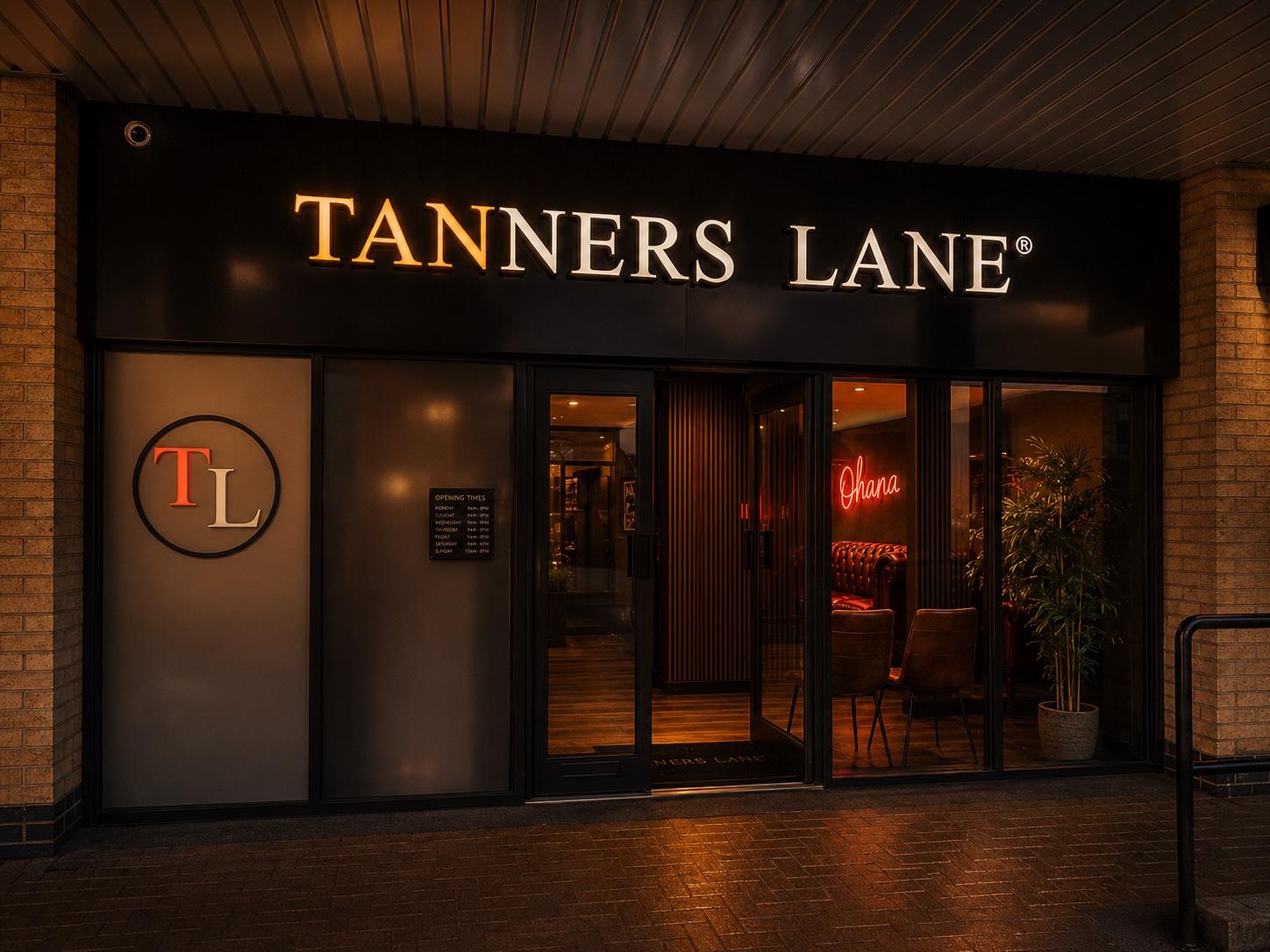 The Tanners Lane storefront at night — the illuminated sign above an open door into the warm, glowing studio